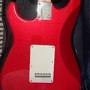 Zadní strana červené elektrické kytary s tvarem typu Stratocaster, umístěná v černém obalu. Na těle je vidět kovová krytka elektroniky a kovový spoj…