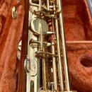 Detailní pohled na část zlaté saxofonu v otevřeném pouzdře s oranžovou výstelkou. Jsou vidět klapy, mechanismus a kovový povrch s drobnými nečistotam…