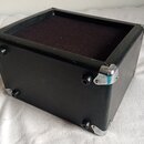 Černý obdélníkový reproduktorový box s černou textilní přední mřížkou. Rohy jsou chráněny kovovými chromovanými zpevňovači, které vykazují mírné oděr…