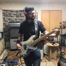 Na fotografii je muž s elektrickou basovou kytarou v bílé barvě s bílým pickguardem. Na hlavě nástroje je viditelné logo Fender. V pozadí stojí kombo…