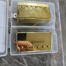 Dvě elektrické kytarové snímače typu humbucker v plastovém průhledném pouzdře. Oba mají zlatý kovový kryt a šest viditelných šroubů. Povrch horního s…