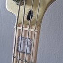 Detail hlavy a horní části hmatníku basové kytary v krémové barvě. Na hlavě je viditelna část černého loga Fender a dva kovové ladičky. Na hmatníku j…