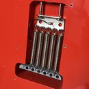 Detailní pohled na červený lakovaný korpus elektrické kytary s vyřezem pro kobylku. V otvoru jsou vidět čtyři kovové pružiny, kovová základna a šesti…