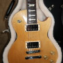 Elektrická kytara typu Les Paul v přírodní dřevěné barvě, umístěná v čalouněném tvrdém kufru. Na hmatníku je viditelný nápis 120th ANNIVERSARY. Kytar…