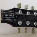 Detail hlavy černé elektrické kytary se značkou Gibson. Na hlavě je bílý nápis Les Paul MODEL a v rámečku text Les Paul STANDARD. Jsou vidět stříbrné…