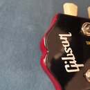 Detail hlavy kytary v černé barvě s červeným okrajem a krémovými ladičkami. Na čelní straně je bílý nápis Gibson a fragment textu DEL. Povrch je glos…