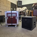 Na fotografii je vidět transportní flightcase v červené barvě s hliníkovými hranami a koly, vedle kterého stojí černý subwoofer. Na flightcasu jsou u…