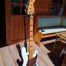 Bílá elektrická basová kytara s jasně hnědým hmatníkem z javoru, umístěná na černém stojanu. Na hlavě je nápis Fender Jazz Bass a logo Fender. Tělo m…