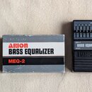 Na fotografii je kytarový pedál ekvalizéru a jeho originální krabička. Černý pedál nese nápisy MEQ-2, BASS EQUALIZER a logo ARION, s ovládacími prvky…