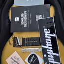 Žlutá elektrická kytara v černém obalu. Na těle kytary a přiložených doplňcích jsou viditelná loga Epiphone a Gibson. Na kytaru jsou položeny reklamn…