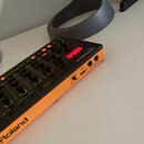 Oranžový sekvenční synthesizer s černým ovládacím panelem. Na přístroji jsou vidět nápisy Roland, TR-8 a označení portů POWER OFF/ON, USB, IN-MIDI-OU…