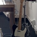 Černá elektrická kytara typu Telecaster se světlým hmatníkem a bílým pickguardem. Na hlavě kytary je vidět nápis Fender. V pozadí jsou umístěny dva č…