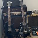 Dvě černé elektrické kytary s tmavým hnedým hmatníkem a černými strapy. Levá kytara má na hlavě nápis Gibson, pravá kytara má na hlavě nápis Ibanez.…