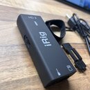 Černý obdélníkový audio rozhraní s nápisy HD 2 a iRig. Zařízení má matný povrch, černý tkaný popruh a vedle něj leží malý plastový černý dílek. Na sp…