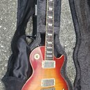 Elektrická kytara typu Les Paul v červeno-žlutém sunburst provedení, umístěná v černém plišovém obalu. Na hlavě hmatníku je bílý nápis Gibson. Kytara…