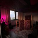 Fotografie zachycuje interiér místnosti s dřevěnou podlahou a kobercem. V prostoru stojí trojdílná akustická clona z dřevěného rámu s červeně vzorova…