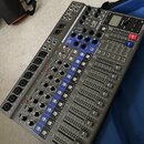 Digitální mixážní pult stříbrné barvy s černým horním panelem. Na zařízení jsou vidět nápisy L-12, L-12 PODKAST a ZOOM. Panel obsahuje řadu posuvníků…