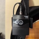 Černý bezdrátový vysílač s nápisem MINI PRO PT 40 a logem AKG. Zařízení je připevněné k černé textilní pásce a má zapojený kabel s konektorem. Povrch…