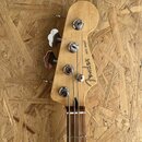 Fotografie zachycuje hlavu basové kytary z přírodního světlého dřeva. Na zadní straně jsou vidět čtyři kovové ladičky a nápisy „Fender“ a „JAZZ BASS®…