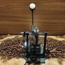 Fotografie znázorňuje mechanismus bicího pedálu v šedé a černé barvě s kovovými součástmi. Zařízení obsahuje vertikální tyč s bílým kulatým koncem, o…