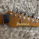 Detail hlavy elektrické kytary z dřeva s přirozenou hnědou barvou. Na dřevě je umístěn bílý nápis Ibanez. Jsou vidět šest kovových ladiček a matice u…