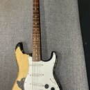 Elektrická kytara typu Stratocaster s černým lakem, který je výrazně odloupaný a odhaluje světlé dřevo korpusu. Na hlavě hmatníku je nápis Fender. Ky…