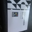 Fotografie zachycuje bílý papírový manuál s černobílým geometrickým vzorem v horní části. Na papíře je uveden nápis DISCOVERER DELAY USER MANUAL a lo…