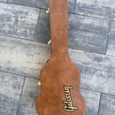 Hnědý tvrdý kufýr na kytaru s texturovaným povrchem. Na spodní části je umístěno žluté logo Gibson. Kufýr má kovové zapínání zlaté barvy.