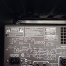Záběr na zadní panel elektronického zařízení s černým povrchem. Na panelu je bílý technický nápis, schéma zapojení a varovné texty začínající slovem…
