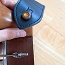 Detailní pohled na hnědý dřevěný buben s chromovaným obručem a ladidly. Na horní části je připevněn černý kožený nebo syntetický popruh s hnědým dřev…