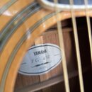 Detailní pohled do otvoru akustické kytary s dřevěným povrchem v žlutohnědé barvě. Uvnitř korpusu je vidět bílá oválná štítková nálepka s nápisy YAMA…
