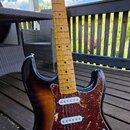 Elektrická kytara typu Stratocaster s barevným přechodem těla od žluté přes hnědou k černé. Nástroj má hnědý hmatník z javorového dřeva a tmřeďový pi…