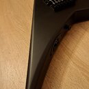 Černá elektrická kytara s matným povrchem a tělem ve tvaru V. Nástroj má dva humbuckery, černý most s pružinami a jeden viditelný ovládací knoflík. N…