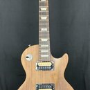 Elektrická kytara typu Les Paul s přírodním dřevěným povrchem a hnědým hnedobělým hmatníkem s trapezoidními vložkami. Na hlavě je nápis Gibson a ozna…