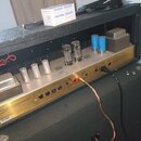 Detail vnitřku kytarového komba s viditelným šasi z anodizovaného hliníku zlatavé barvy. Na šasi jsou osazeny čtyři elektronky a dva modré kondenzáto…
