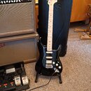 Černá elektrická kytara typu Stratocaster s bílým hmatníkem a bílým pickguardem, opřená o černý tvrdý kufřík. Na hlavě kytary je nápis Fender STRATOC…