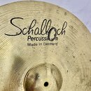 Detailní pohled na mosazný činel s koncentrickým žebrováním povrchu. Na povrchu je černý nápis „Schalloch“, pod ním slovo „Percussion“ a text „Made i…