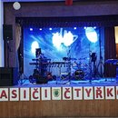 Fotografie zachycuje hudební sestavu na pódiu s modrým osvětlením a pozadím. Je zde vidět bicí souprava s logem na basovém bubnu, klávesy a několik r…