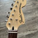 Fotografie zachycuje hlavu elektrické kytary z přírodního světlého dřeva s hnedým hmatníkem. Na povrchu je černý nápis Fender® STRATOCASTER® a v horn…