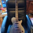 Elektrická kytara typu Les Paul s tmavě hnědým průsvitným lakem a viditnou strukturou dřeva. Nástroj má dva humbuckery, čtyři ovládací knofíky a jede…