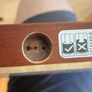 Detail dřevěného dílu v hnědé barvě s vyvrtaným otvorem. Uvnitř otvoru jsou vidět dva malé šrouby a stopy po lepidle nebo opravě. Vedle otvoru je nal…