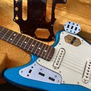 Modře třpytivá elektrická kytara s bílým pickguardem a hnedým hmatníkem. Vedle kytary leží samostatný tmavě červený pickguard s texturou tortise. Na…