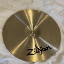 Zlatavý mosazný činel s jemným kruhovým broušením povrchu. Na spodní části je černý nápis Zildjian. U středu činelu je viditelná bílá papírová nálepk…