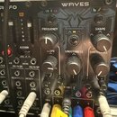 Černý modulární syntetizátor s nápisem WAVES. Na panelu jsou ovládací prvky s popisky FREQUENCY, SLOPE, SMOOTHNESS, SHAPE a SHIFT/LEVEL, dále značení…