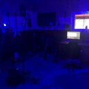 Fotografie zachycuje interiér hudebního studia v modrém osvětlení. V místnosti jsou vidět dvě elektrické kytary, jedna z nich je bílá s špičatým tvar…