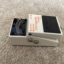 Bílý kytarový ladič značky BOSS, model TU-3 Chromatic Tuner. Zařízení má bílé kovové tělo s černým pedalem a analogovým budíkovým displejem. Na boku…