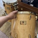 Dva bubny v přírodním dřevěném provedení s gold hardwarem. Na předním bubnu je viditelný nápis Pearl Masters Custom Maple. V pozadí je druhý buben s…