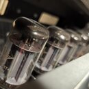 Detailní pohled do vnitřku přístroje, kde je vidět řada skleněných elektronických lamp. Na přední lampě je viditelný nápis "ECC83" a logo znáčky. Pov…