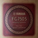 Detailní záběr na štítek uvnitř akustické kytary. Štítky je červený s okroužkovým ornamentem a obsahuje nápisy YAMAHA, FG750S, Made in China a YAMAHA…