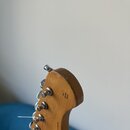 Detail zadní strany hlavy elektrické kytary z přírodního dřeva. Na povrchu je vidět nápis "Fine Tuned Sound" a svisle orientovaný text "V7-Series". H…